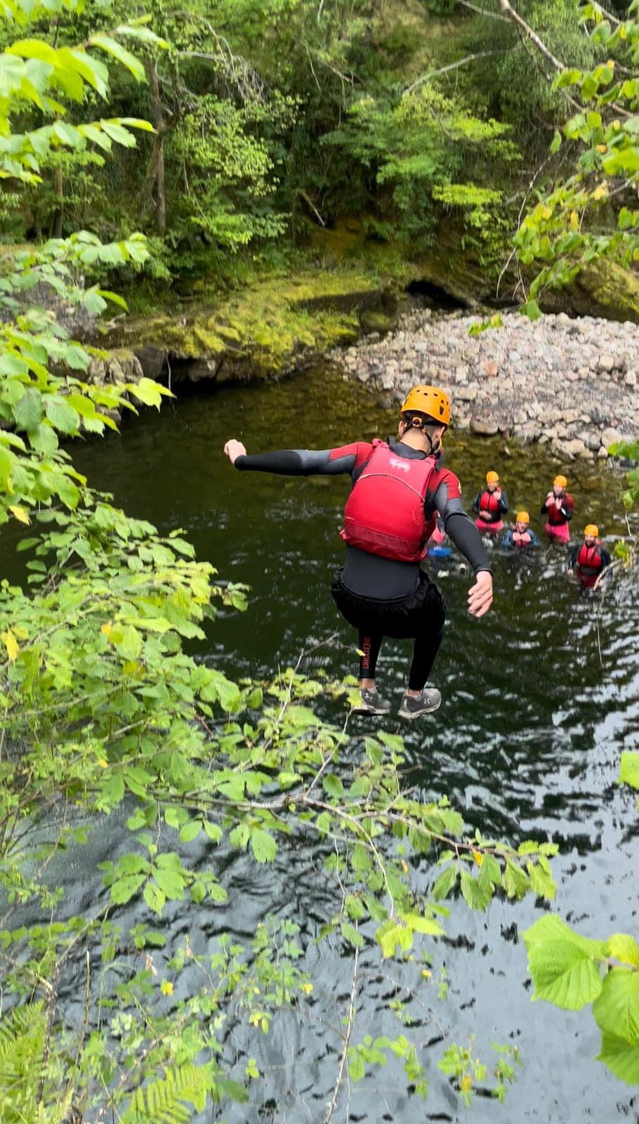 A person in a canyoning trip in Wales is jumping off a waterfall while companions are waiting at the bottom.